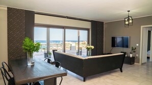 Una sala de estar moderna con sofá, mesa de comedor, sillas, televisor y un balcón con vista al mar.