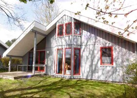 Une maison moderne avec un toit en pente, de grandes fenêtres aux montures rouges et une véranda bien visible.