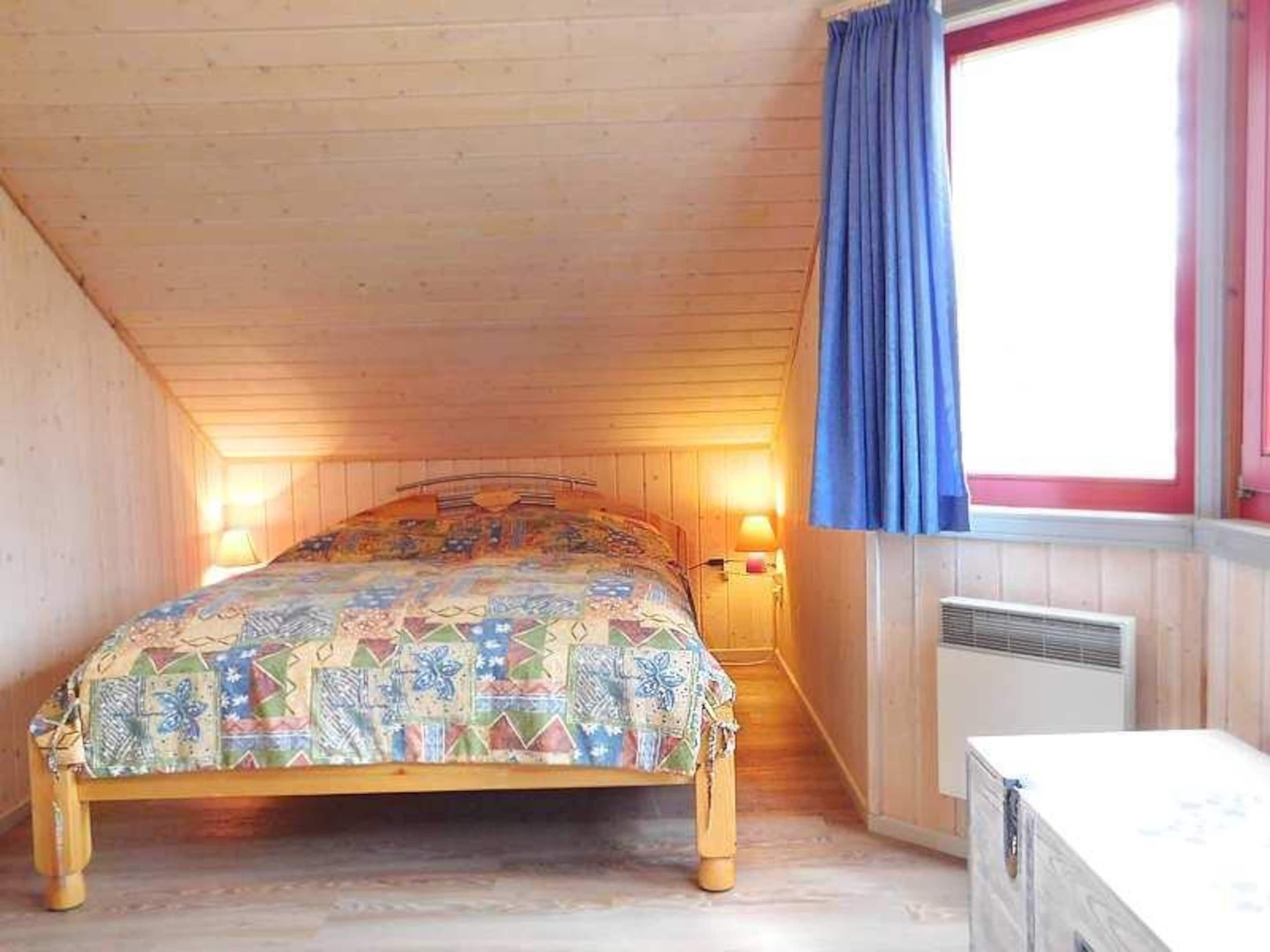 Een slaapkamer met een houten plafond, een bed met een kleurrijke sprei, een raam met blauwe gordijnen en een wandradiator.