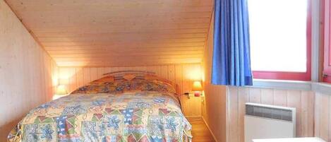 Een slaapkamer met een houten plafond, een bed met een kleurrijke sprei, een raam met blauwe gordijnen en een wandradiator.