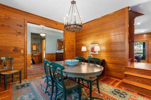 A wooden-paneled room with a round table, chairs, a chandelier, and a patterned rug. - Windows on the Water (Pawleys Island)