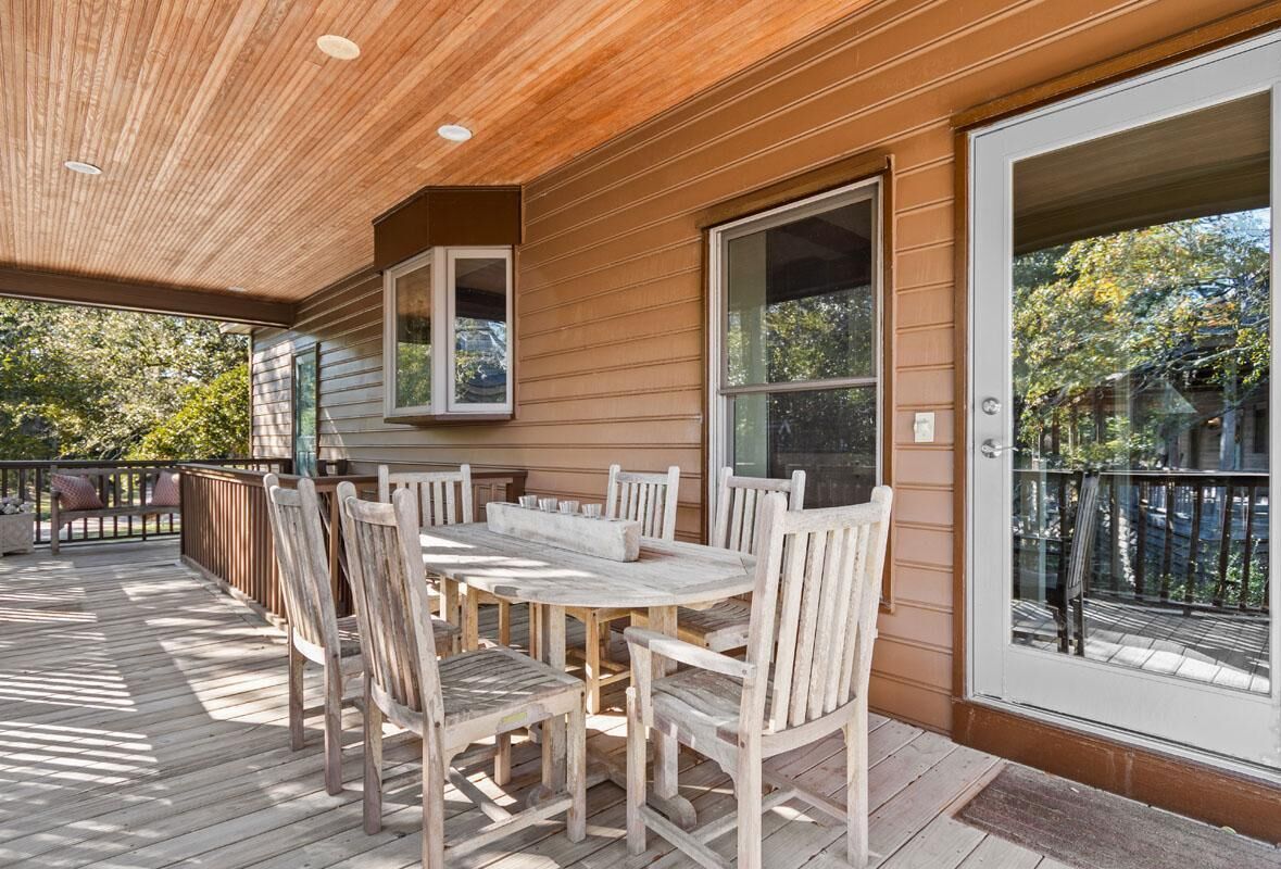 Una terraza de madera con mesa y sillas, ventanas amplias y vista a árboles.