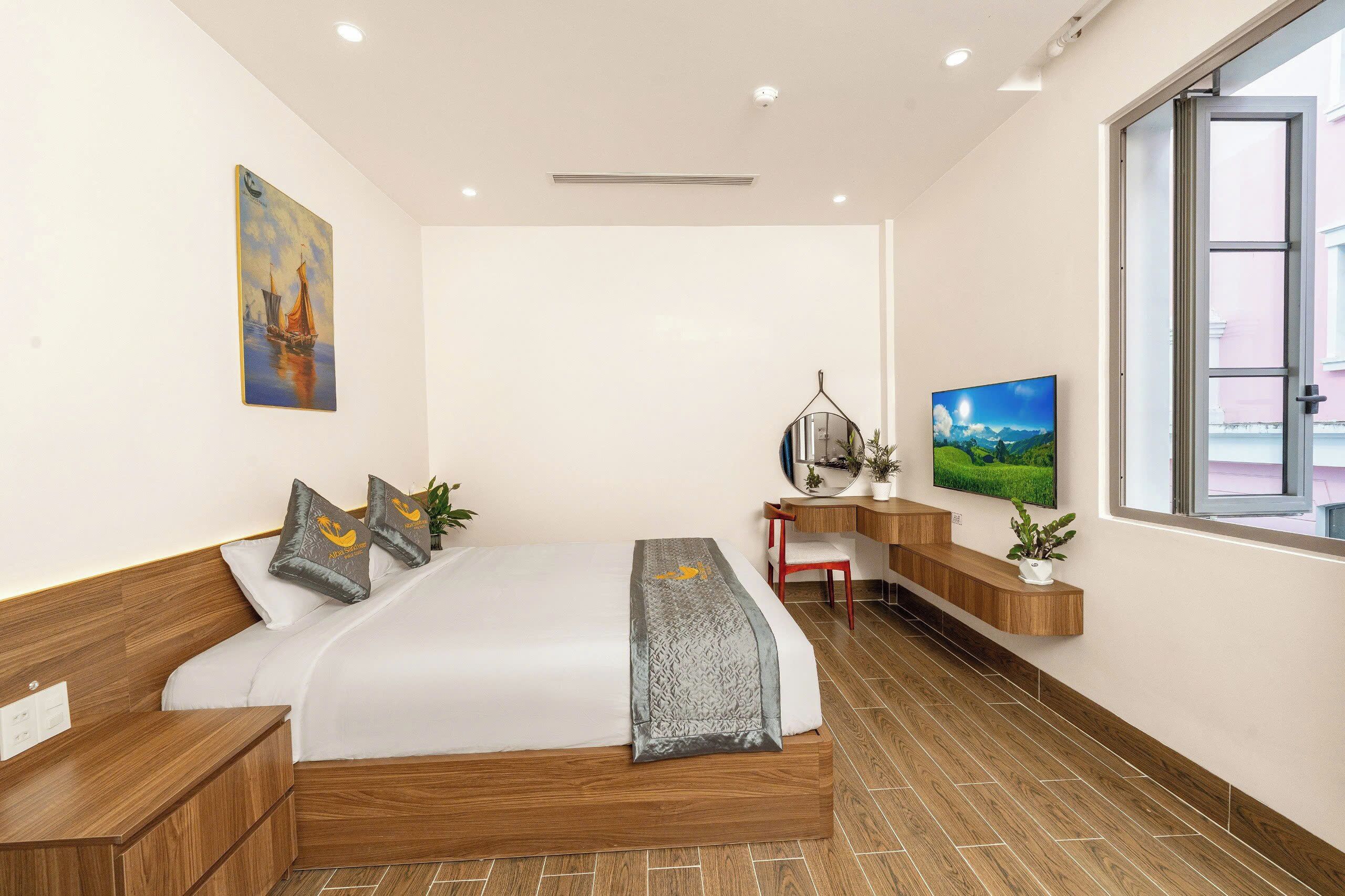 A modern hotel room with a large bed, wooden headboard, a desk with a chair, a TV, and a window with a city view.