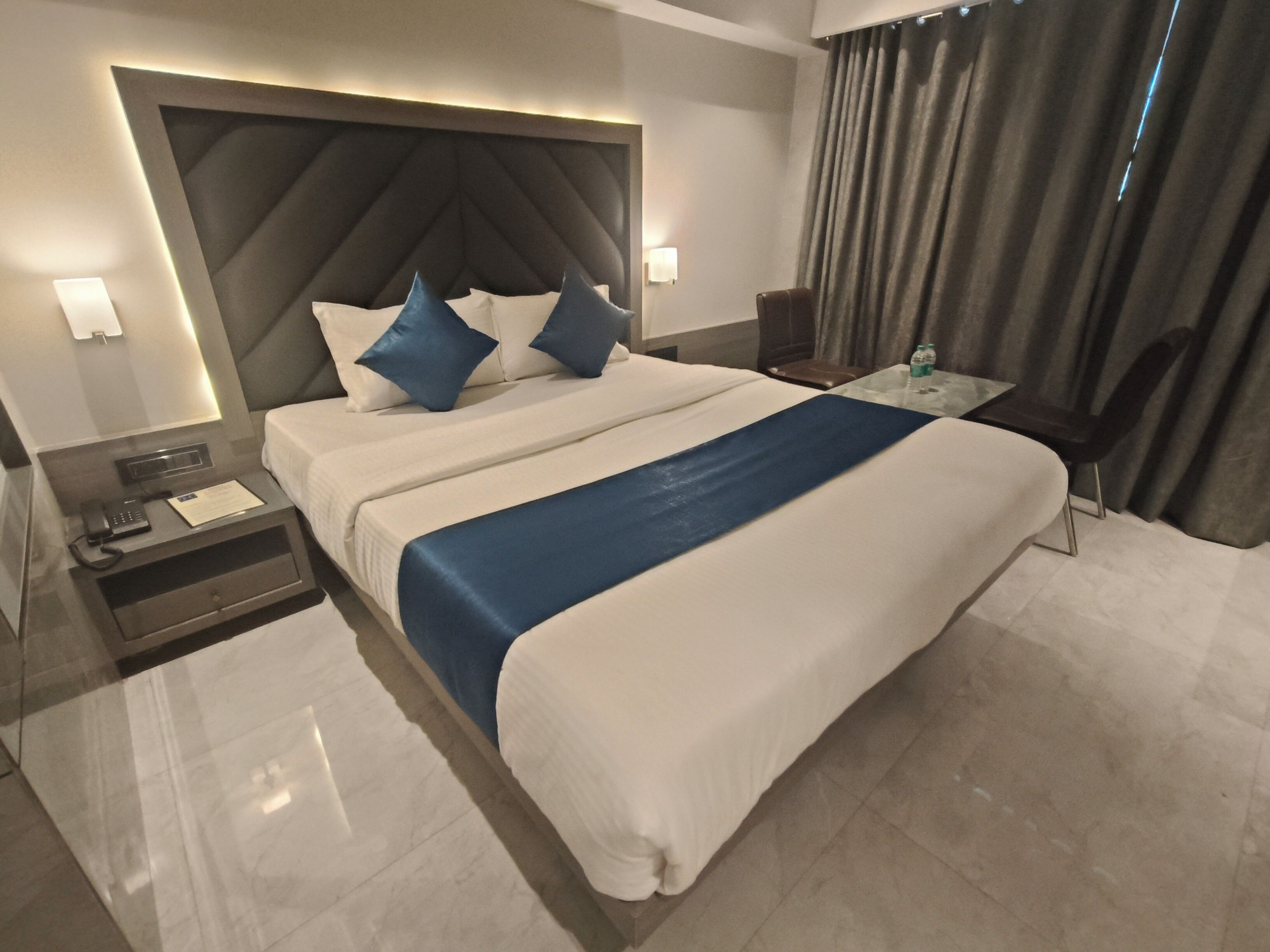 Quarto de hotel moderno com uma cama grande, mesa de cabeceira com telefone e papéis, uma pequena mesa com uma garrafa e uma cadeira.