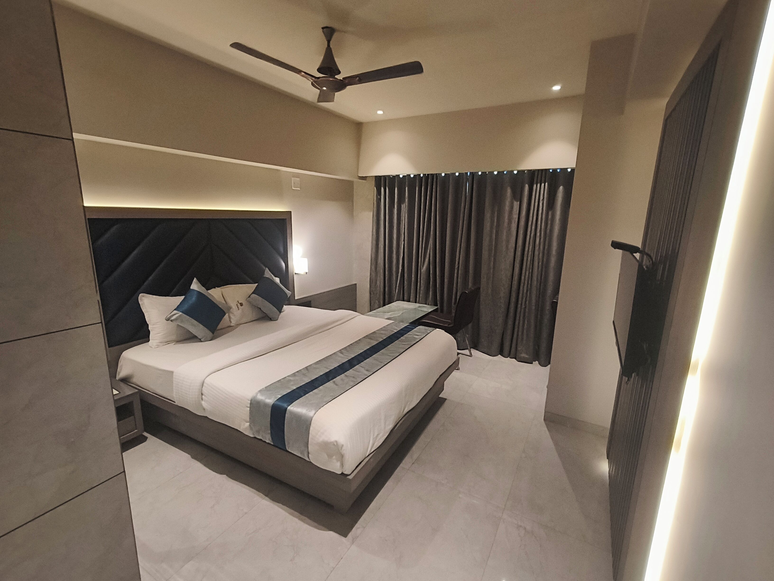 A modern hotel room with a large bed, a ceiling fan, and a view of the city through the window.