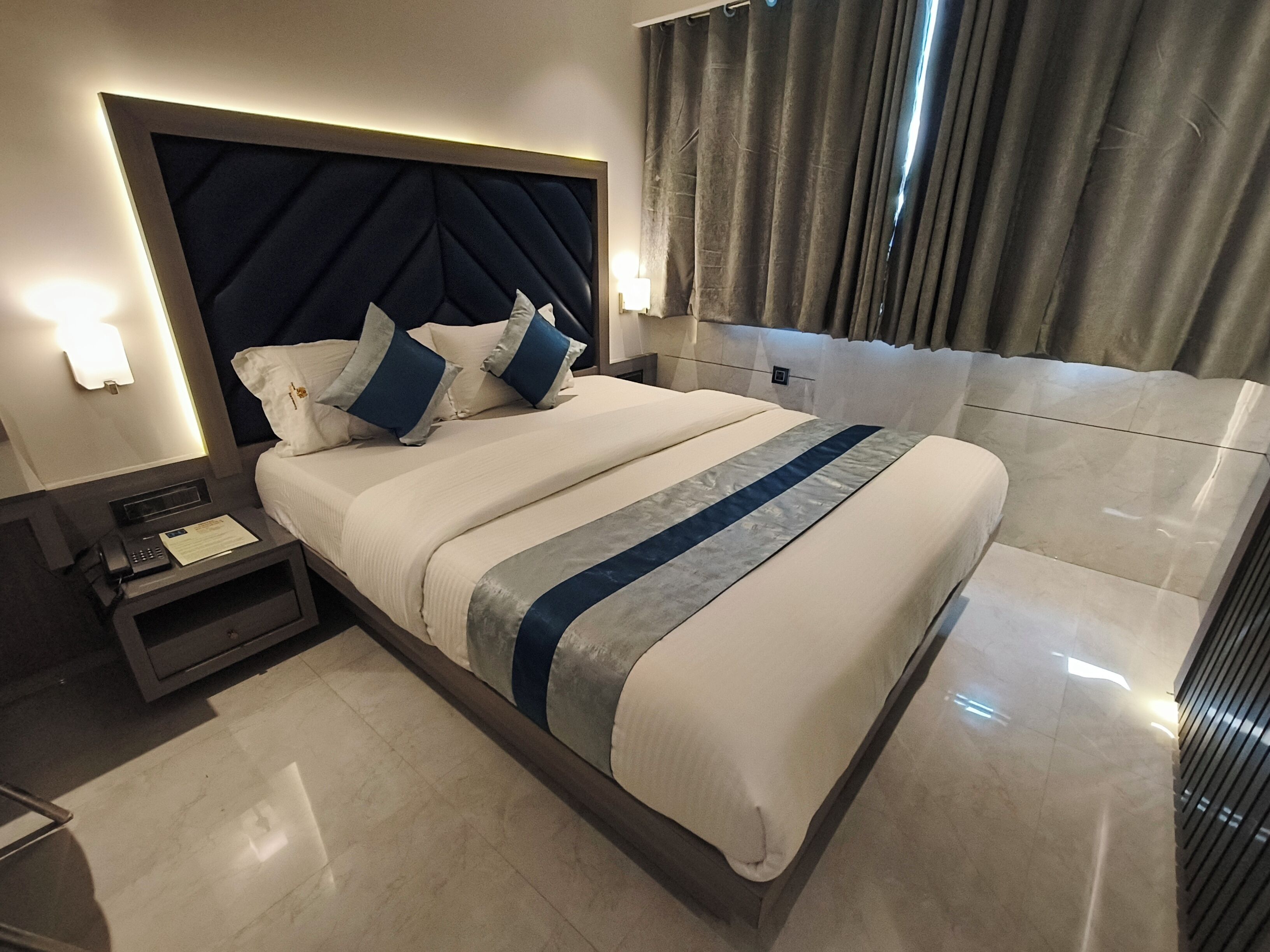 Una habitación de hotel moderna con una cama grande, una mesita de noche con un teléfono y documentos, y un cabecero con una lámpara.