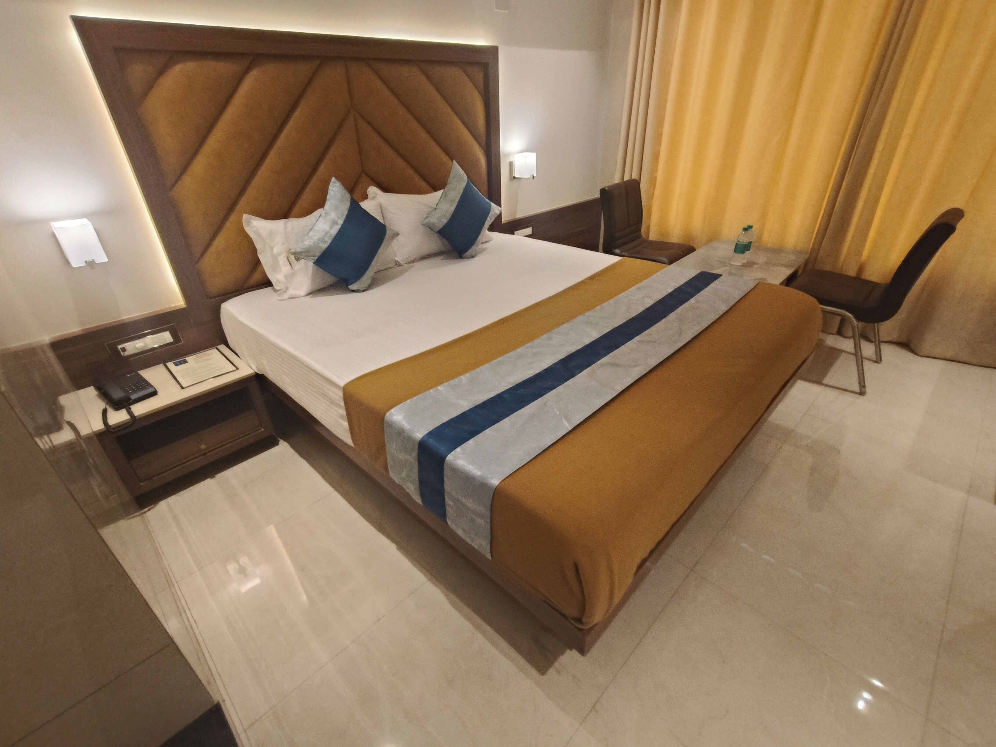 A hotel room with a large bed, a bedside table with a phone and a notepad, a chair, a small table with a bottle, and a curtain.