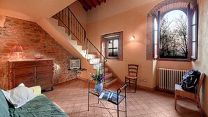 A cozy living room with a stone wall, a wooden staircase, a green sofa, a coffee table with a vase, and a window with a view of trees. - Holiday Home in Reggello (Reggello)