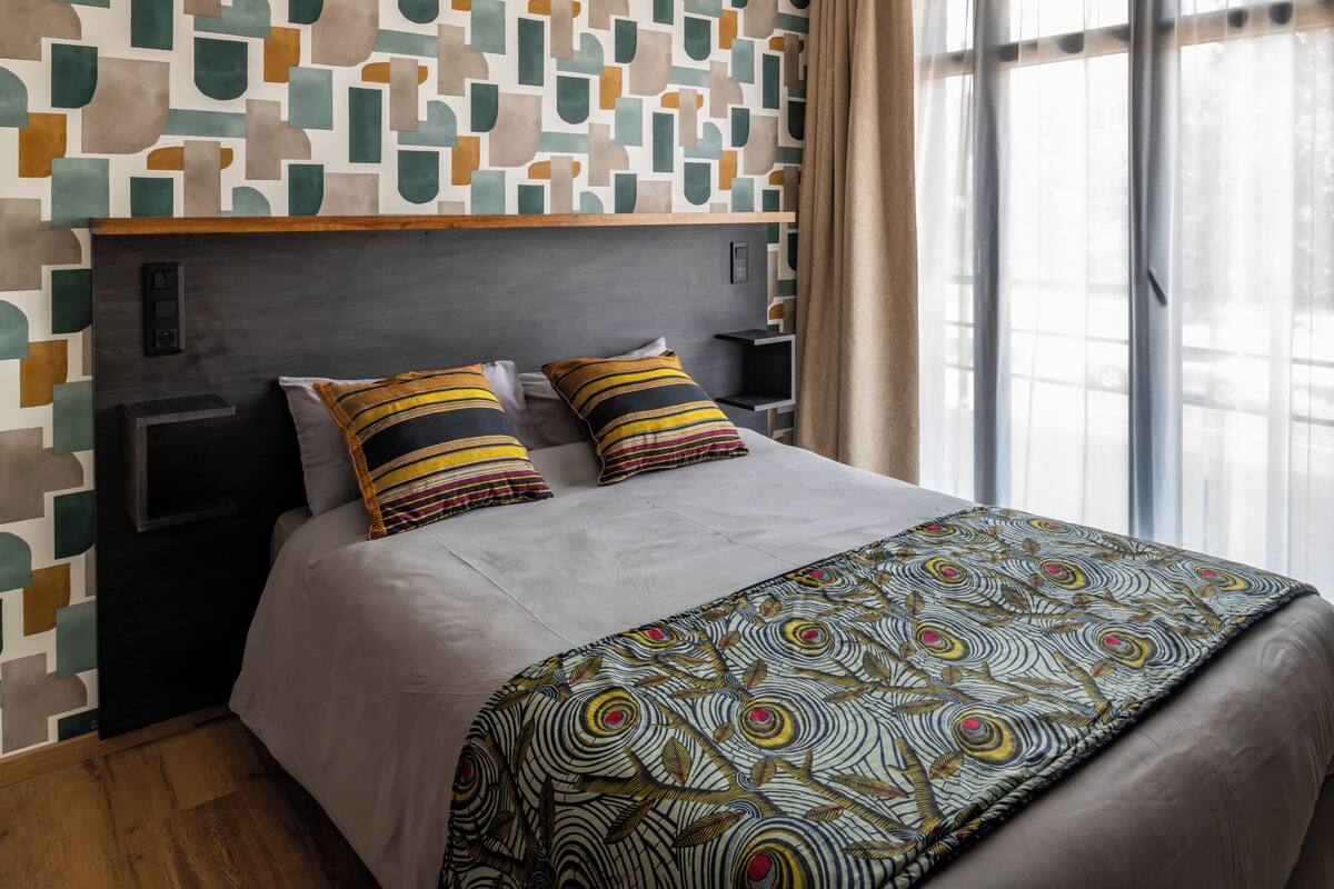 A hotel room with a modern geometric-patterned wall, a large bed with striped pillows, and a patterned throw blanket.