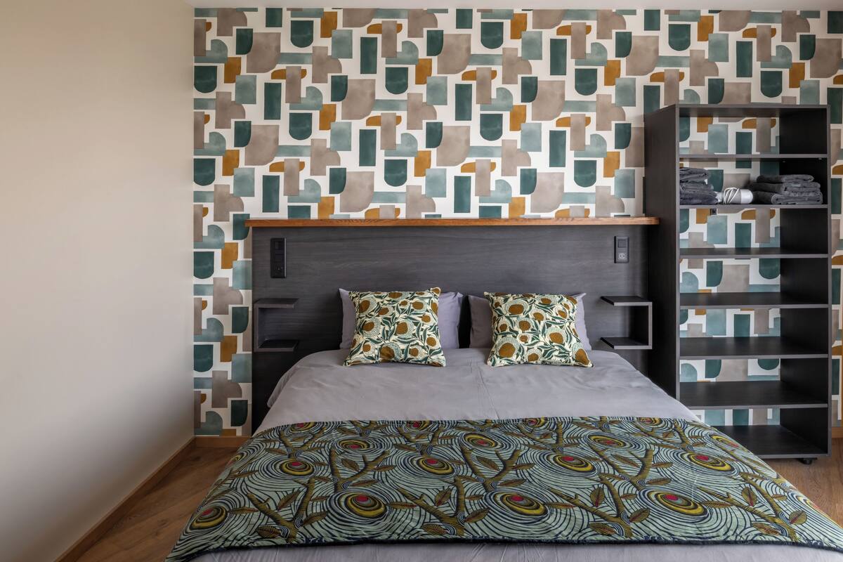 A modern bedroom with a geometric patterned wall, a dark headboard, a grey bedspread, and a patterned throw blanket.