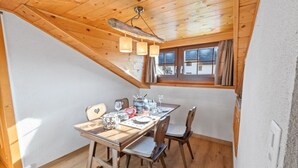 A dining area with a wooden table set for four, a window with curtains, and a sloped ceiling with hanging lights. - Montana Village - appartement rénové, 5 pers (Montana Village)