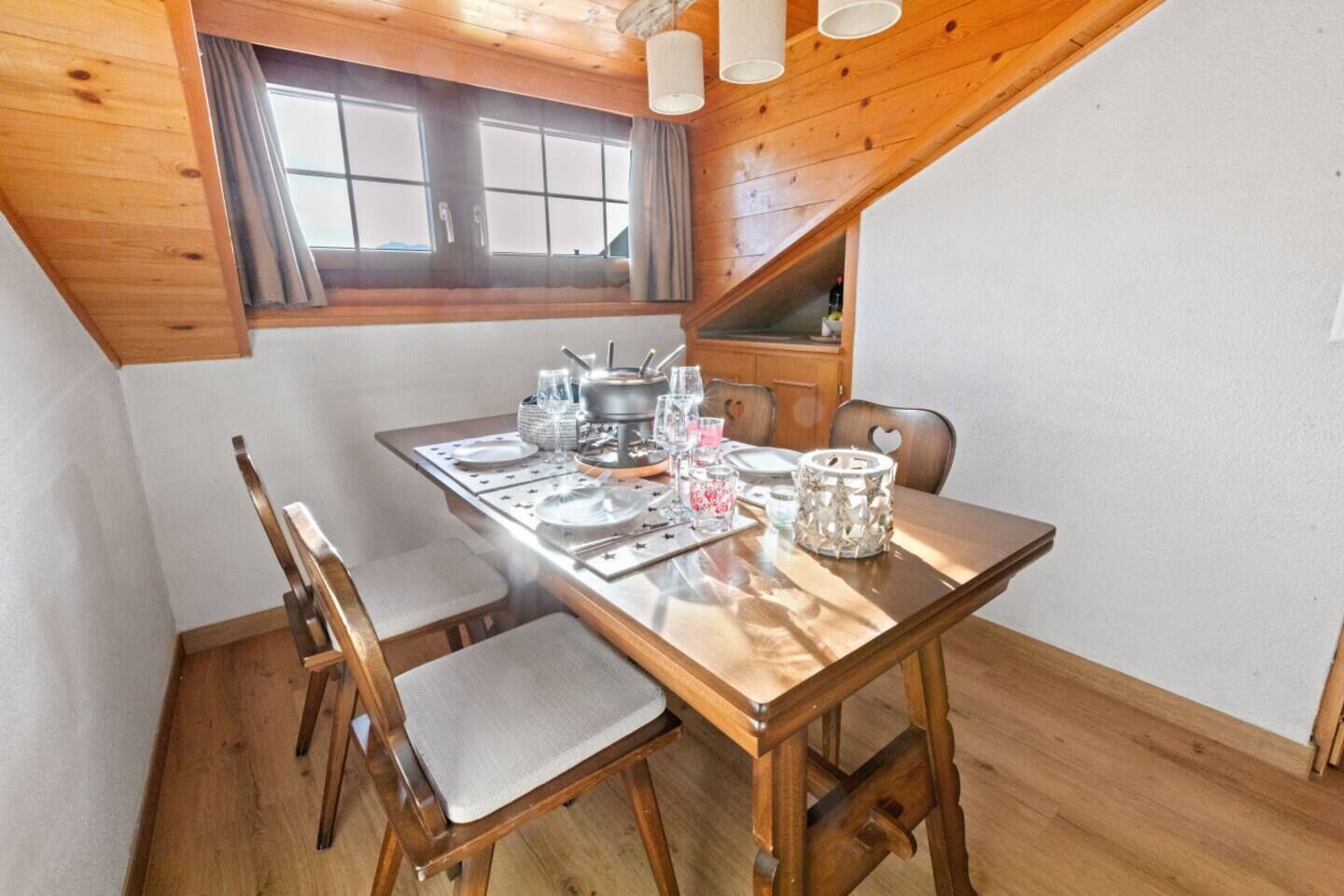 A dining area with a wooden table set for four, a window above, and a sloped ceiling.