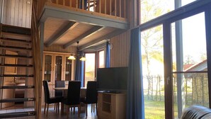 Ett rymligt vardagsrum med träinredning, ett matbord med stolar, en tv och stora fönster som ger utsikt över omgivningarna.
