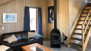 Ett mysigt rum med en svart lädersoffa, ett träbord, en svart vedkamin, ett fönster med blå gardiner och en trätrappa.