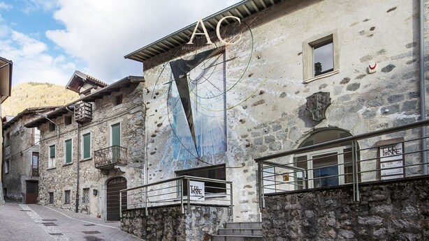A narrow street with stone buildings, one with a mural and the letters "AC" on the facade.