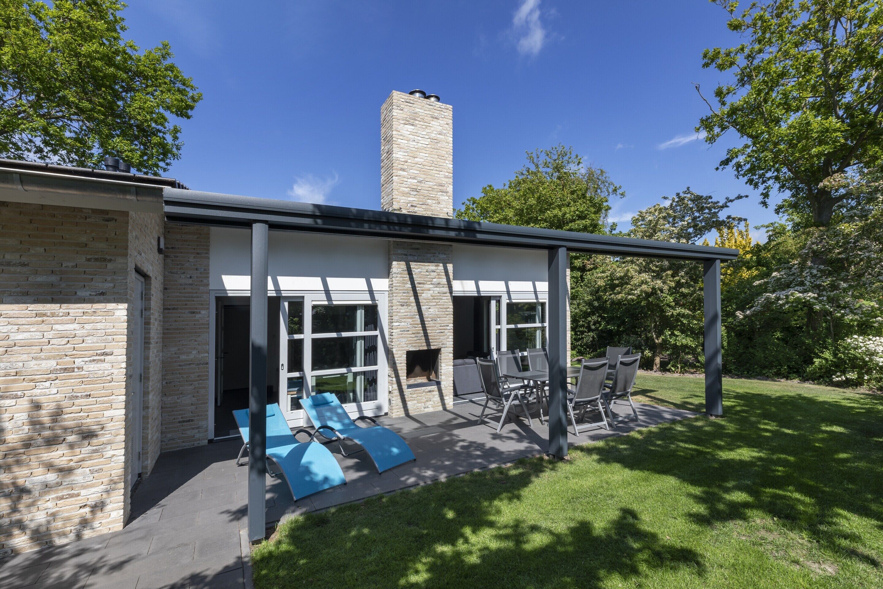 Et moderne hus med murstensfacade, skorsten, en overdækket terrasse med udendørs møbler og en græsplæne.