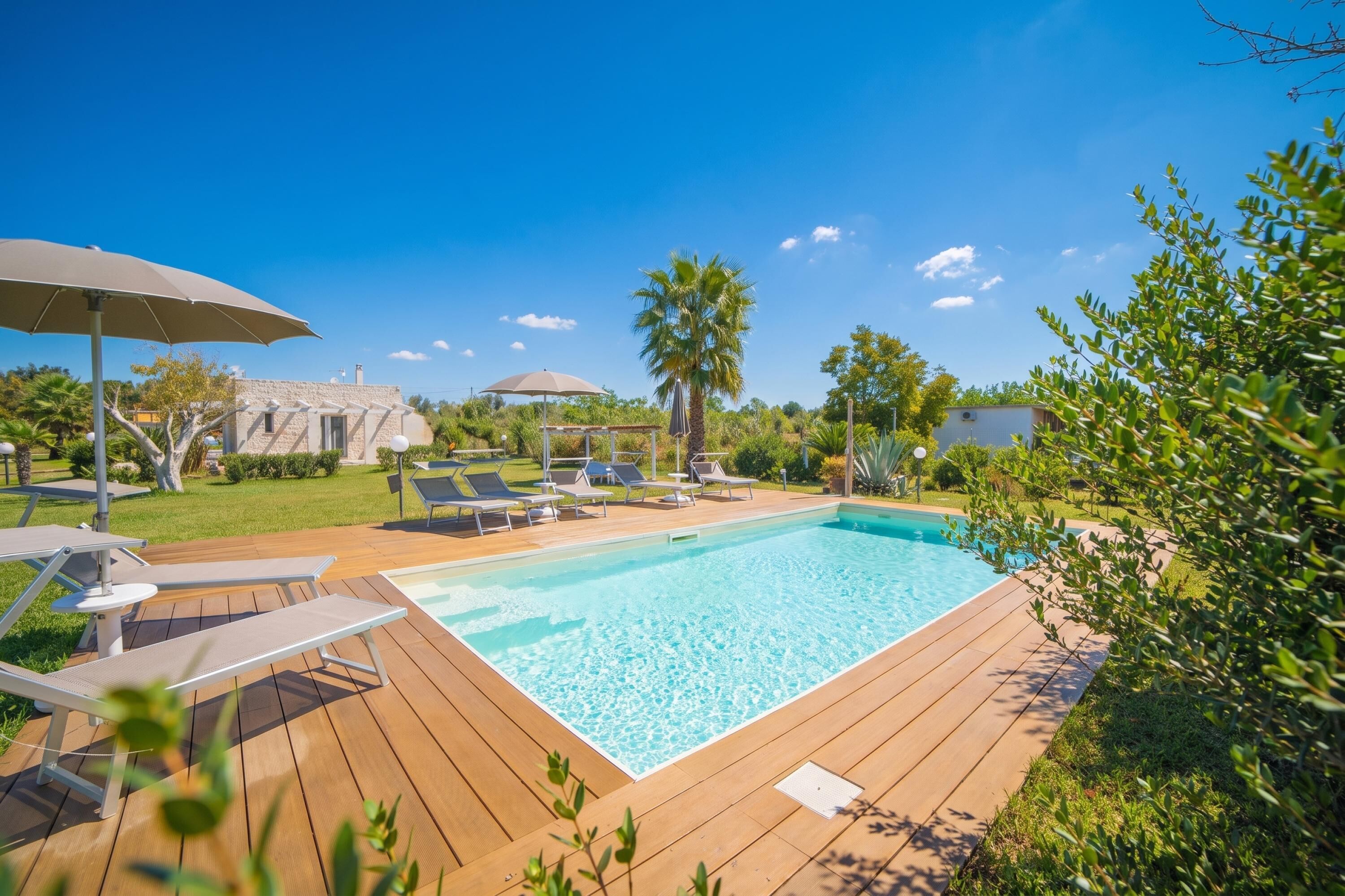 A swimming pool with lounge chairs and umbrellas, surrounded by greenery and a clear sky.