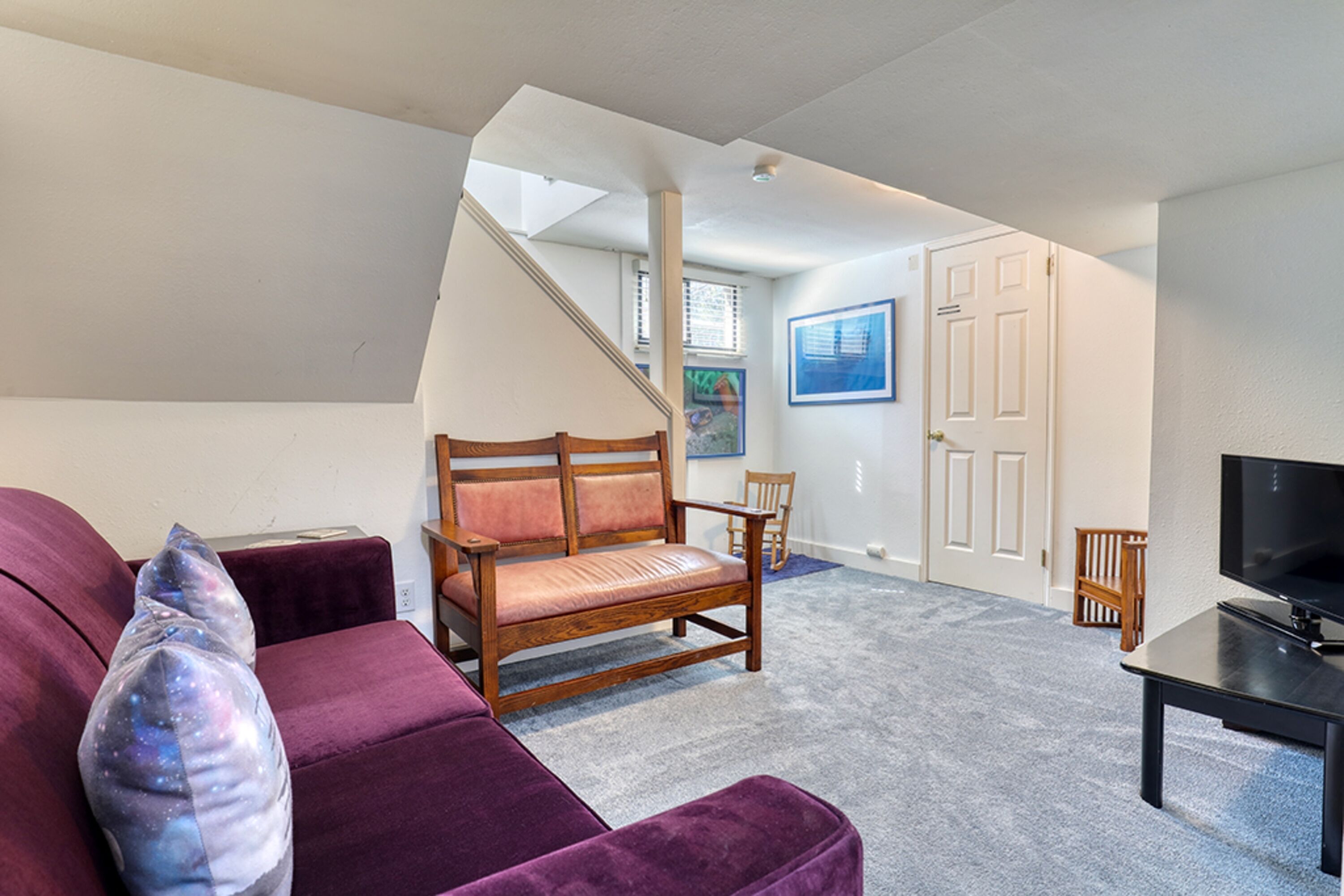 Een woonkamer met een paarse bank, een houten stoel, een televisie op een standaard en een trap die naar een andere kamer leidt.
