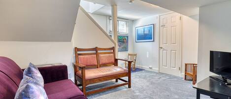 Een woonkamer met een paarse bank, een houten stoel, een televisie op een standaard en een trap die naar een andere kamer leidt.