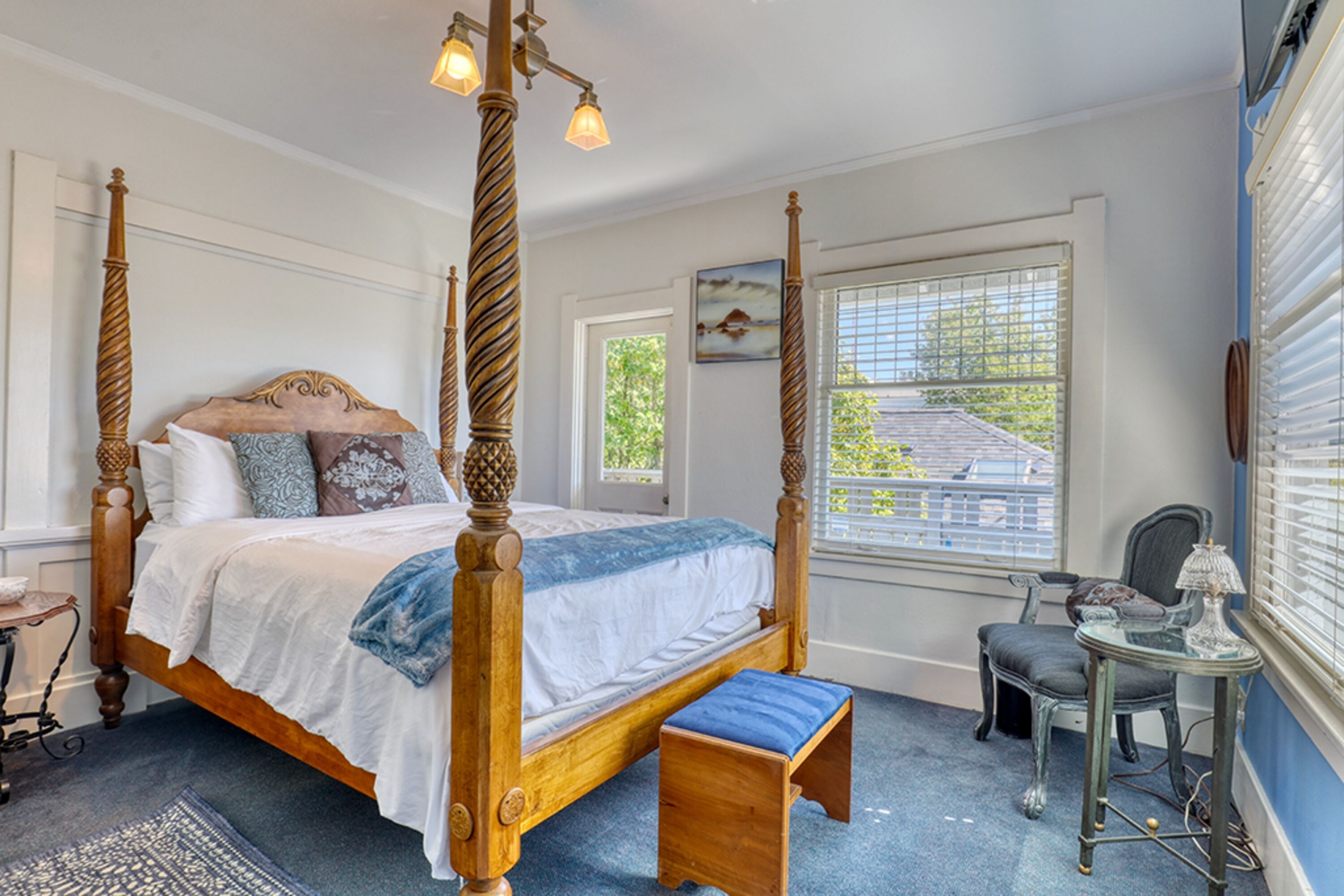 A four-poster bed with a wooden frame and a bench at the foot, a small table with a lamp, and a window with blinds.