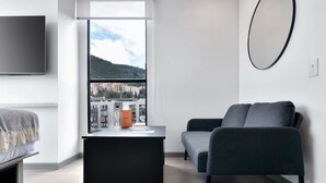 Ein modernes Wohnzimmer mit einem schwarzen Schreibtisch, einem grauen Sofa, einem Wand-Fernseher und einem großen Fenster mit Blick auf die Stadt.