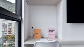 Ein schlicht eingerichtetes Zimmer mit Schreibtisch, einer Flasche, einem Willkommensschild und Blick auf Gebäude und Bäume.