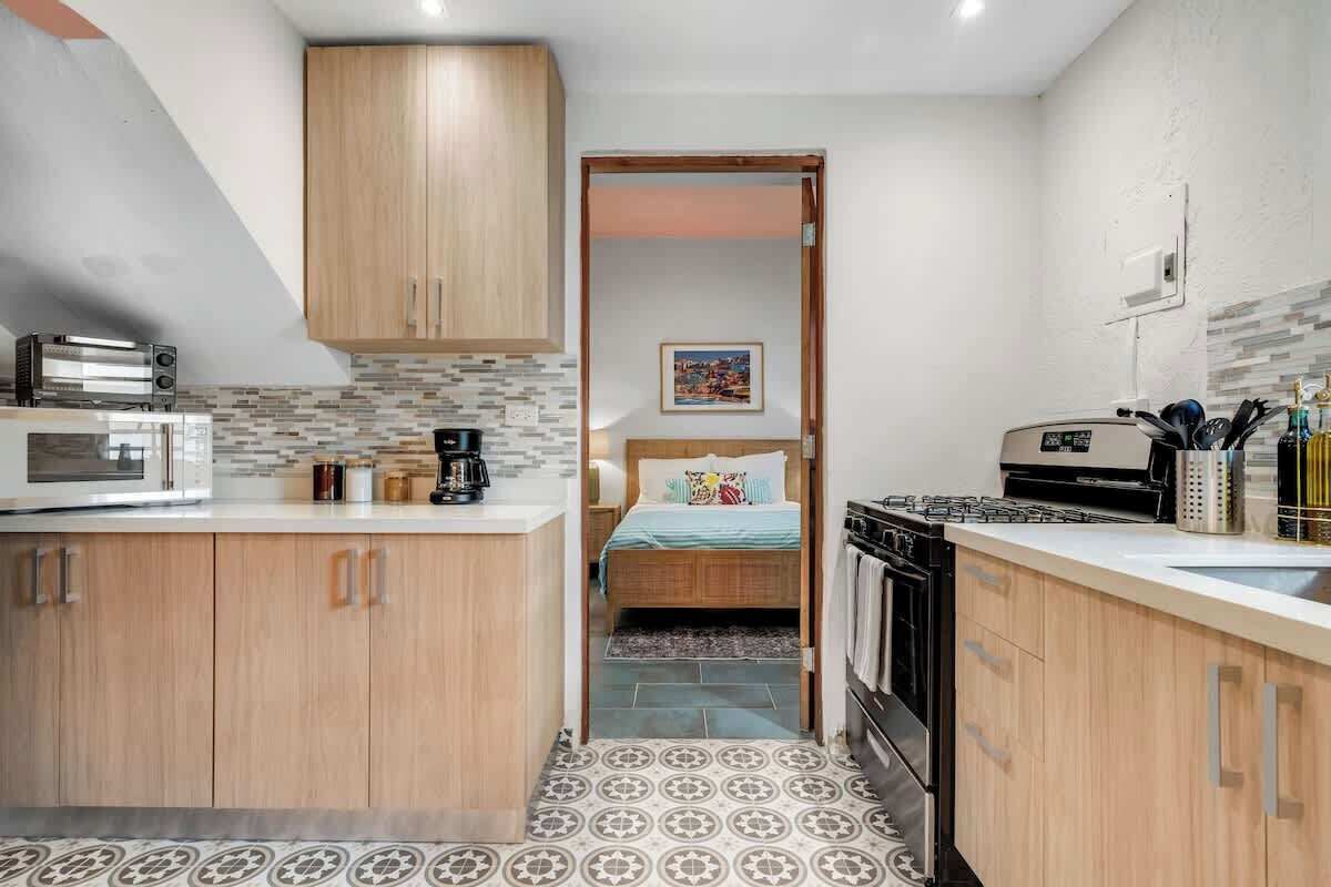Une cuisine compacte avec des meubles en bois, un four à micro-ondes, une cuisinière et un évier. On aperçoit, au fond, un lit recouvert d’une couverture à rayures bleues et blanches.