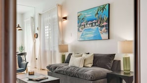 A modern living room with a grey sofa, a coffee table, and two lamps. There is a painting of a tropical scene on the wall.