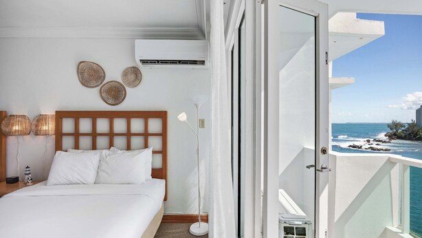 Una habitació d'hotel amb un llit, una taula de nit, aire condicionat fixat a la paret i vista a l'oceà des d'un balcó.