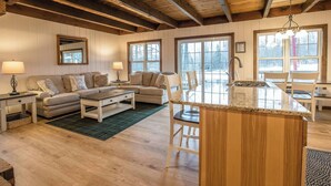 A cozy living room with a wooden ceiling, a sofa set, a dining table, and a kitchen island.