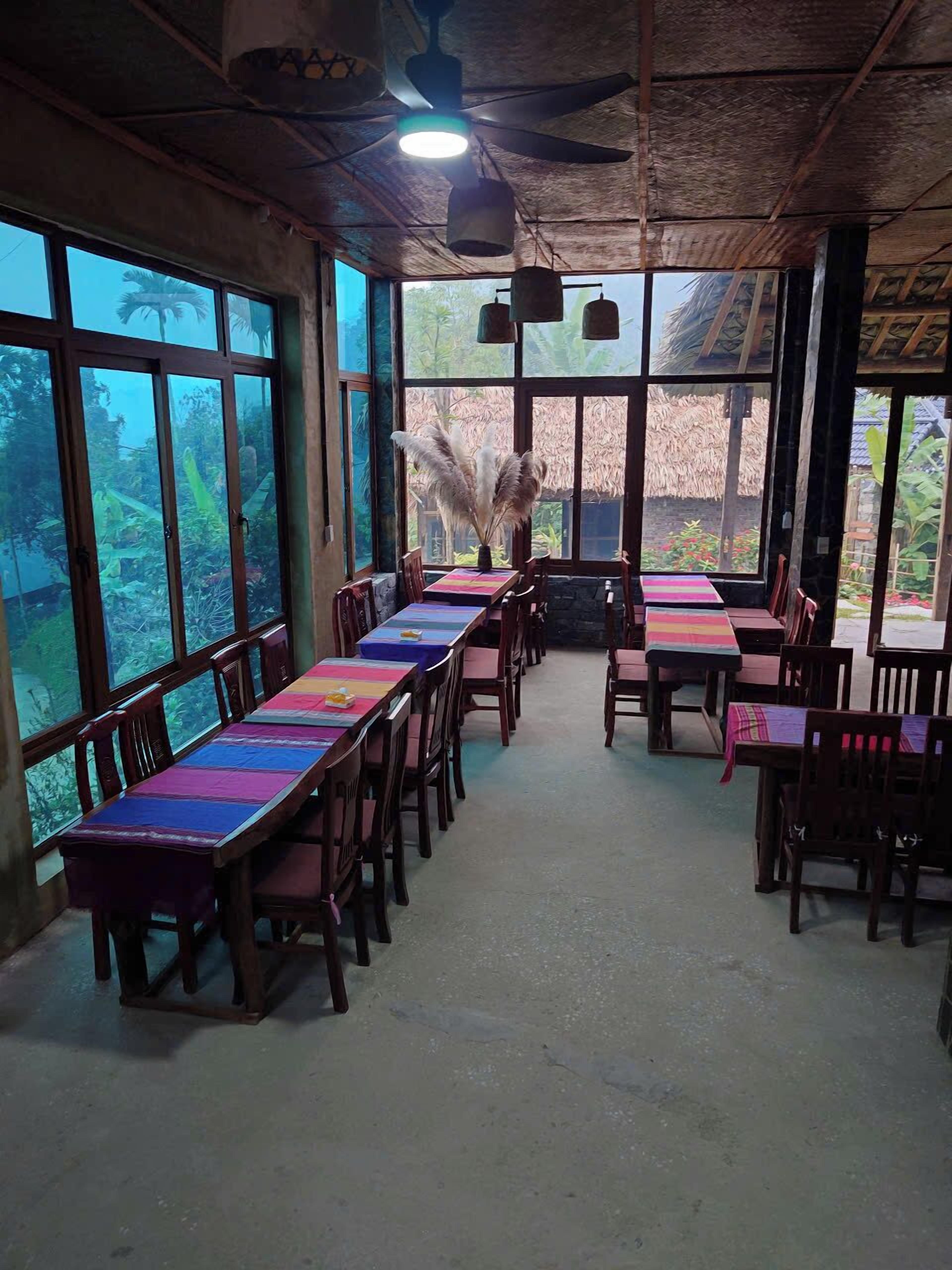 A restaurant with long wooden tables and chairs, large windows, and a ceiling fan.