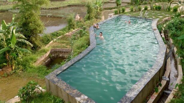 Piscina com borda de pedra, rodeada por vegetação exuberante e campos em socalcos ao fundo.