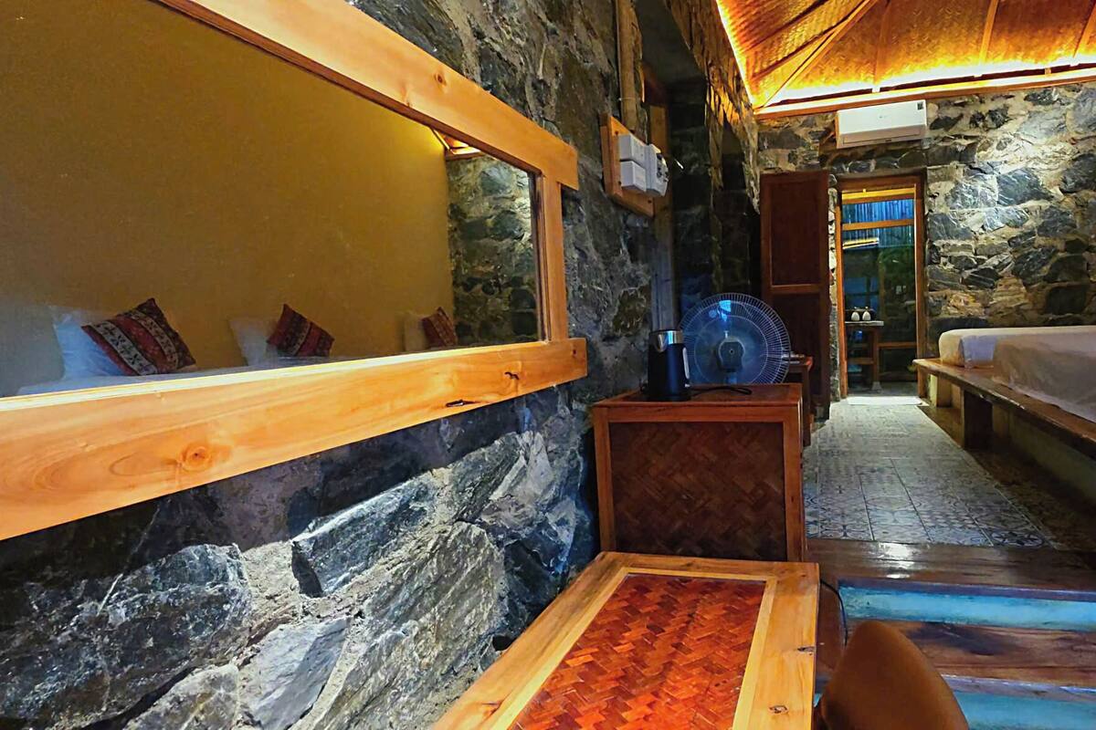 A cozy room with a stone wall, wooden beams, a bed, a small table, and a fan.