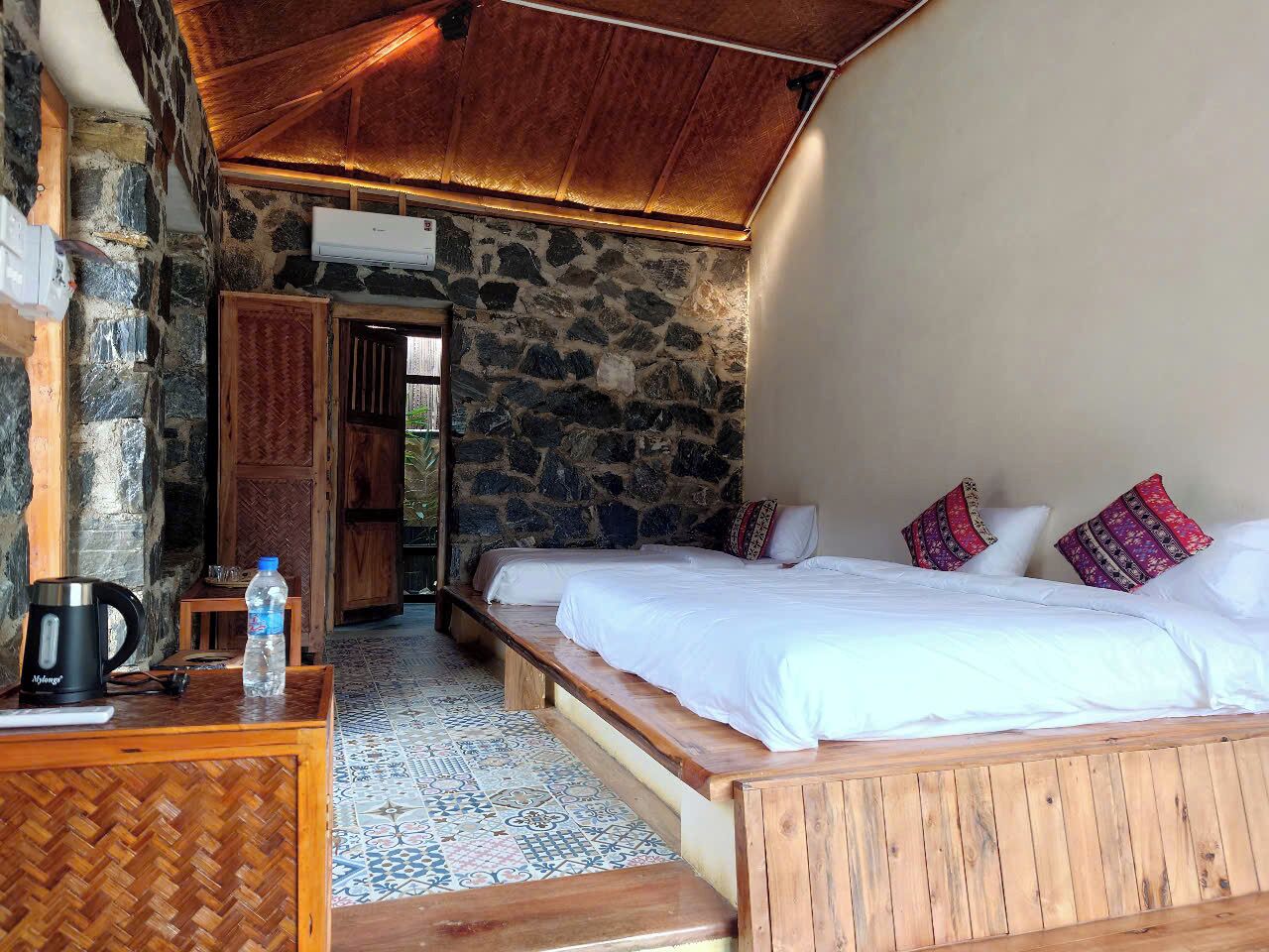 Quarto aconchegante com parede de pedra, cama de madeira e pequena mesa com chaleira e garrafa de água.