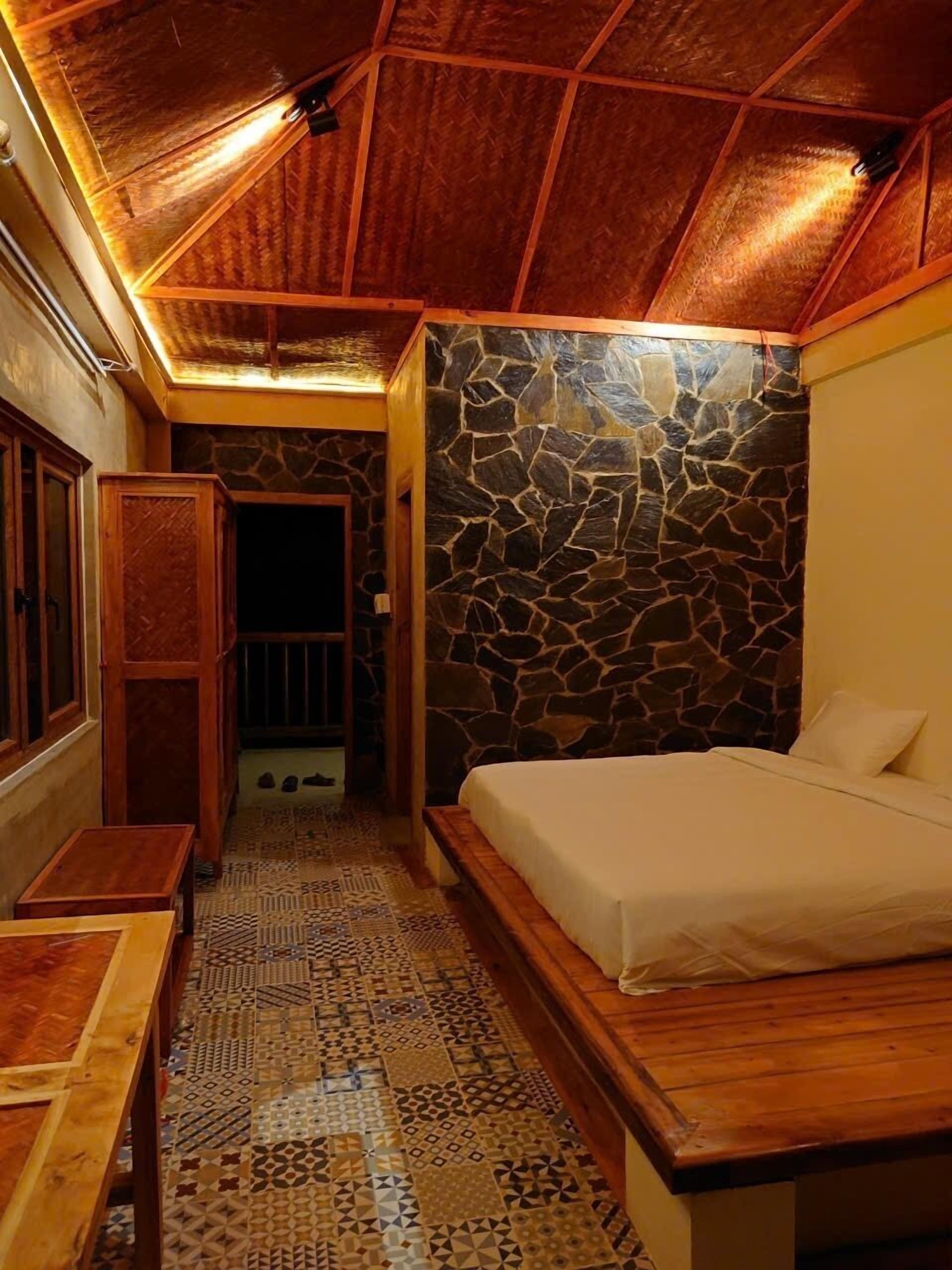 A bedroom with a stone wall, a bed, a wooden bench, and a patterned floor.