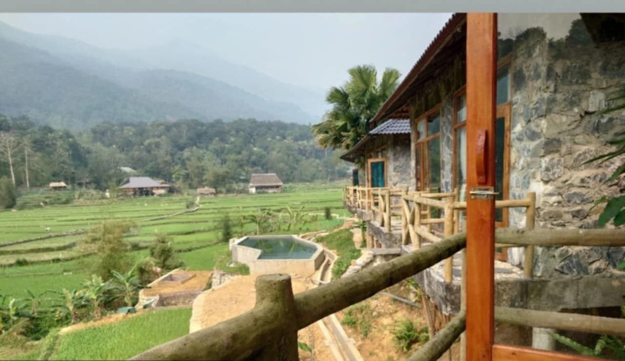 A rustic bungalow with a wooden balcony overlooking terraced fields and a mountain backdrop.