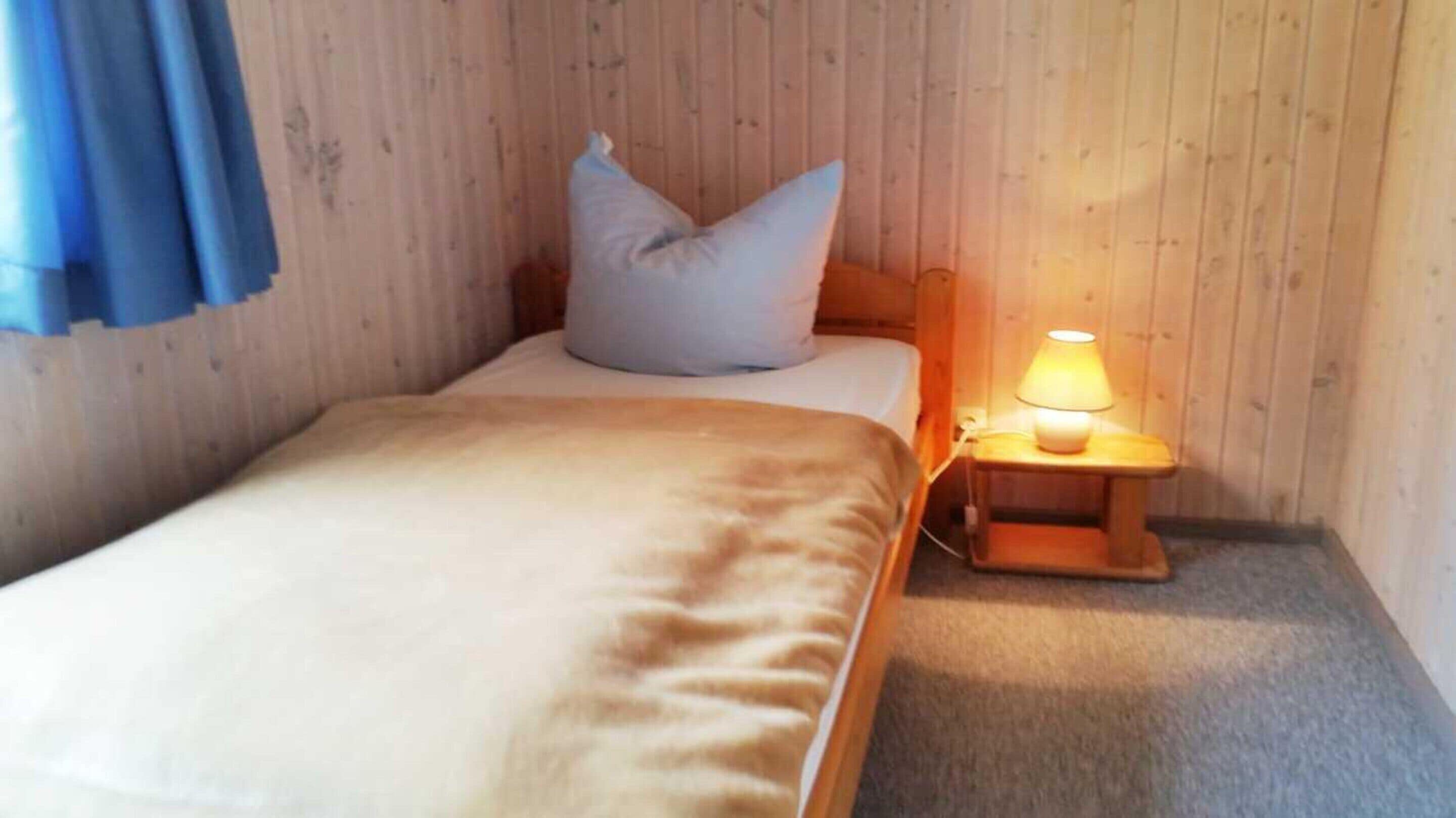Una cama individual con un almohadón blanco y una colcha color arena en una habitación con paredes de madera y suelo alfombrado.