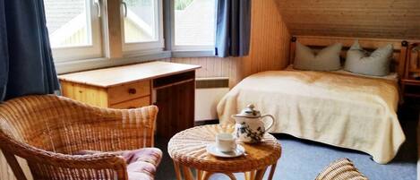 Una habitación acogedora con una cama, un sillón de mimbre, una mesita con tetera y una ventana con cortinas.