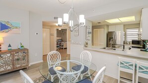 Un comedor con mesa de vidrio y sillas blancas, una araña, una cocina con refrigerador de acero inoxidable, y un pasillo que conduce a otra habitación.
