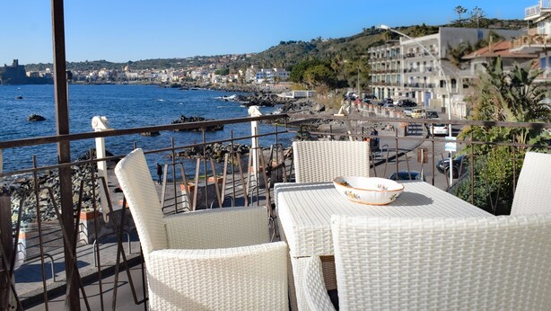 A balcony with wicker chairs and a table, overlooking a coastal town with buildings and a rocky shoreline. - Wiew Acitrezza (Acicastello)