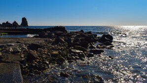A rocky shoreline with waves crashing against the rocks and a clear blue sky. - Wiew Acitrezza (Acicastello)