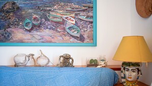 A room with a blue patterned sofa, a painting of boats on a beach, and a lamp with a face on the nightstand. - Wiew Acitrezza (Acicastello)