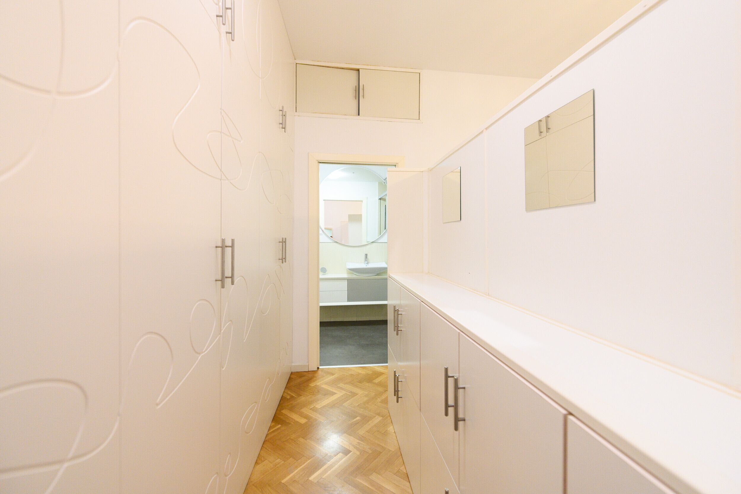 A narrow hallway with white walls and wooden flooring, leading to a bathroom with a sink and mirror.