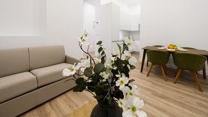 Ett modernt vardagsrum med en beige soffa, en matplats med ett träbord och gröna stolar samt en vas med vita blommor.