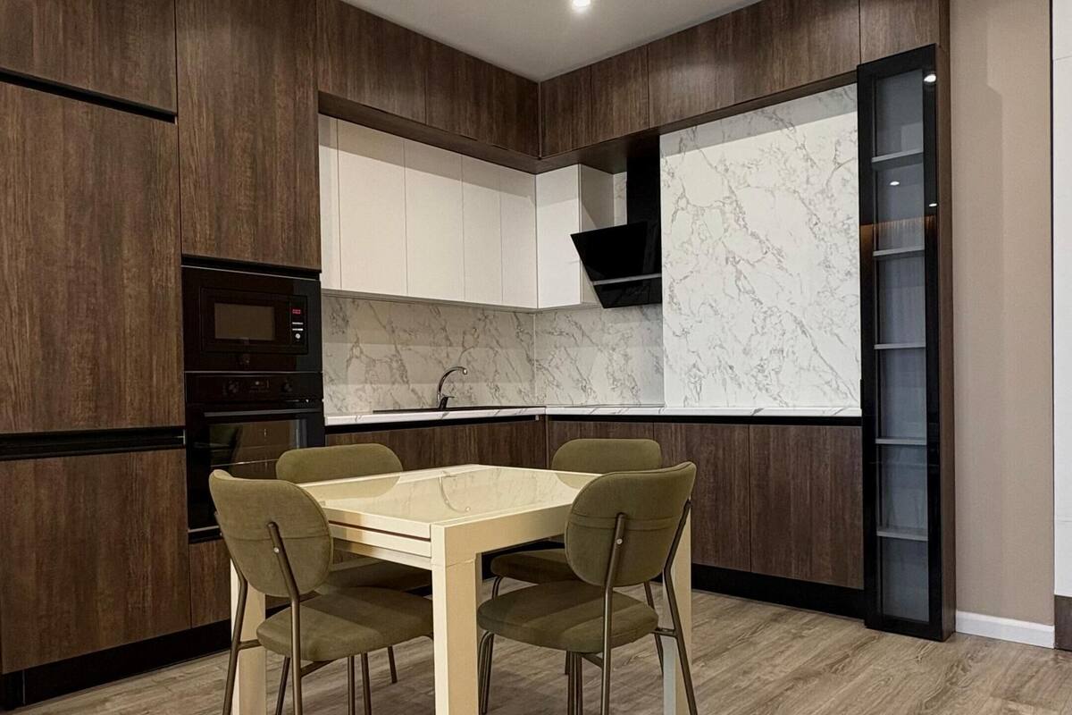 Cozinha moderna com armários em madeira escura, painel de parede em mármore e área de jantar com mesa e cadeiras de madeira.
