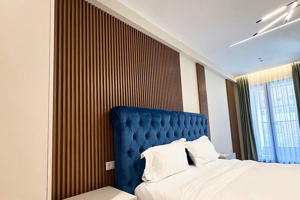 Quarto moderno com cabeceira estofada azul, colcha branca e painel de parede de madeira.