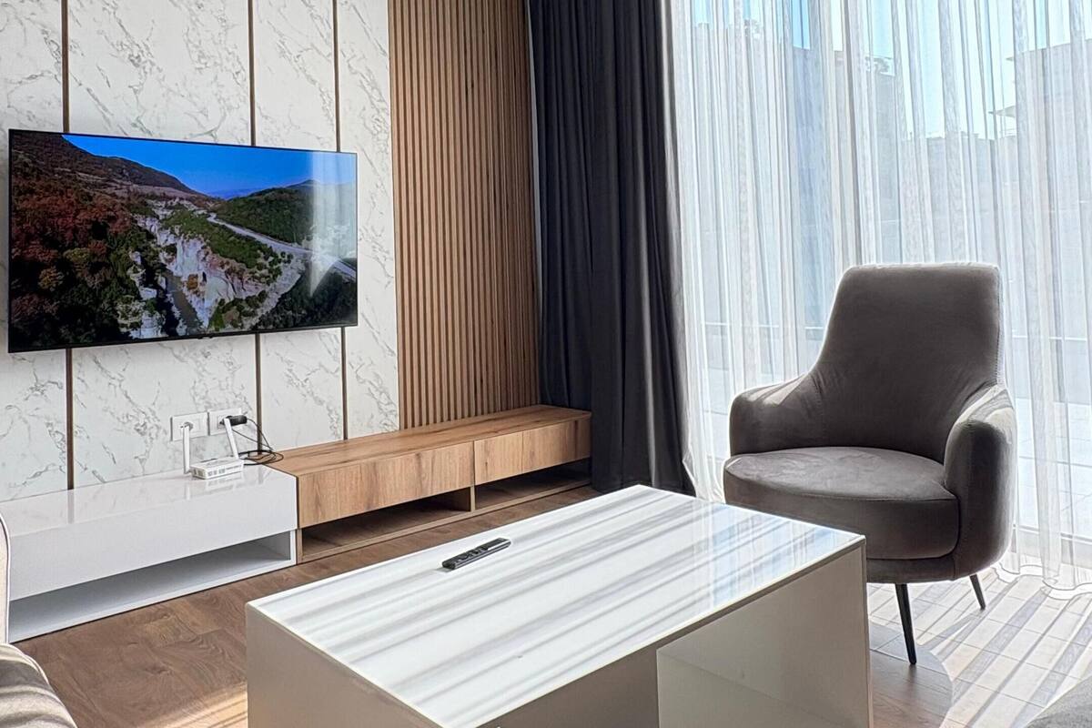 Uma sala de estar moderna com uma TV de tela plana fixada em uma parede de mármore, uma mesa de centro branca, uma poltrona cinza e um lustre preto no teto.
