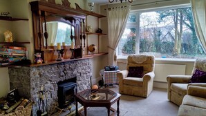A cozy living room with a stone fireplace, a wooden mantel, and a large window with a view of trees.