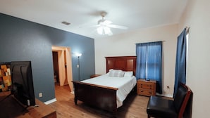 Ein Schlafzimmer mit einem Bett, einem Sessel, einem Schreibtisch, einem Deckenventilator und einem Fenster mit Vorhängen.