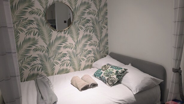 A neatly made bed with two rolled towels and a pillow, a round mirror on the wall, and a patterned wallpaper. - Cosy Coastal Apartment in Lytham St Anne's (Lytham St. Anne's)